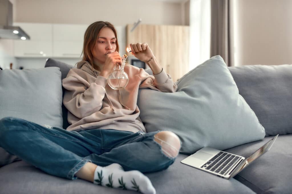 Young caucasian woman sitting on the couch at home and lighting cannabis in the bowl of glass water pipe or bong. Cannabis and weed legalization concept. Horizontal shot