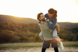 Smiling man carrying a woman on his back, laughing and looking at each other during an active outdoor moment at dusk
