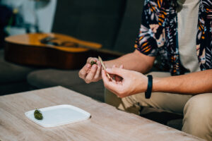 Asian young man sitting on a couch with a cannabis bud in his hand and smoking a joint relaxing time at home. A guitar is in the background