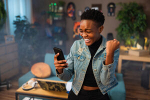 Happy african american teen girl lady woman with short hair using cell phone holding in hands smiling celebrating raising fists.