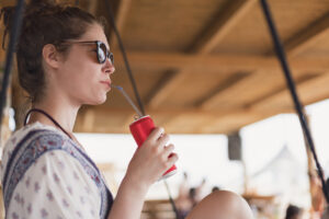 Attractive young woman relaxing on the swing at the beach bar and drinking soda.