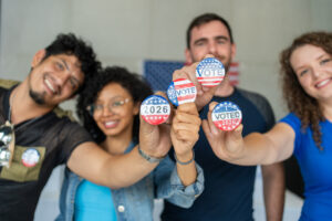 diverse group of smiling friends holding 2026 Vote election I voted buttons or pins at a US election polling station. In the background, american flag at the background, democracy in America USA